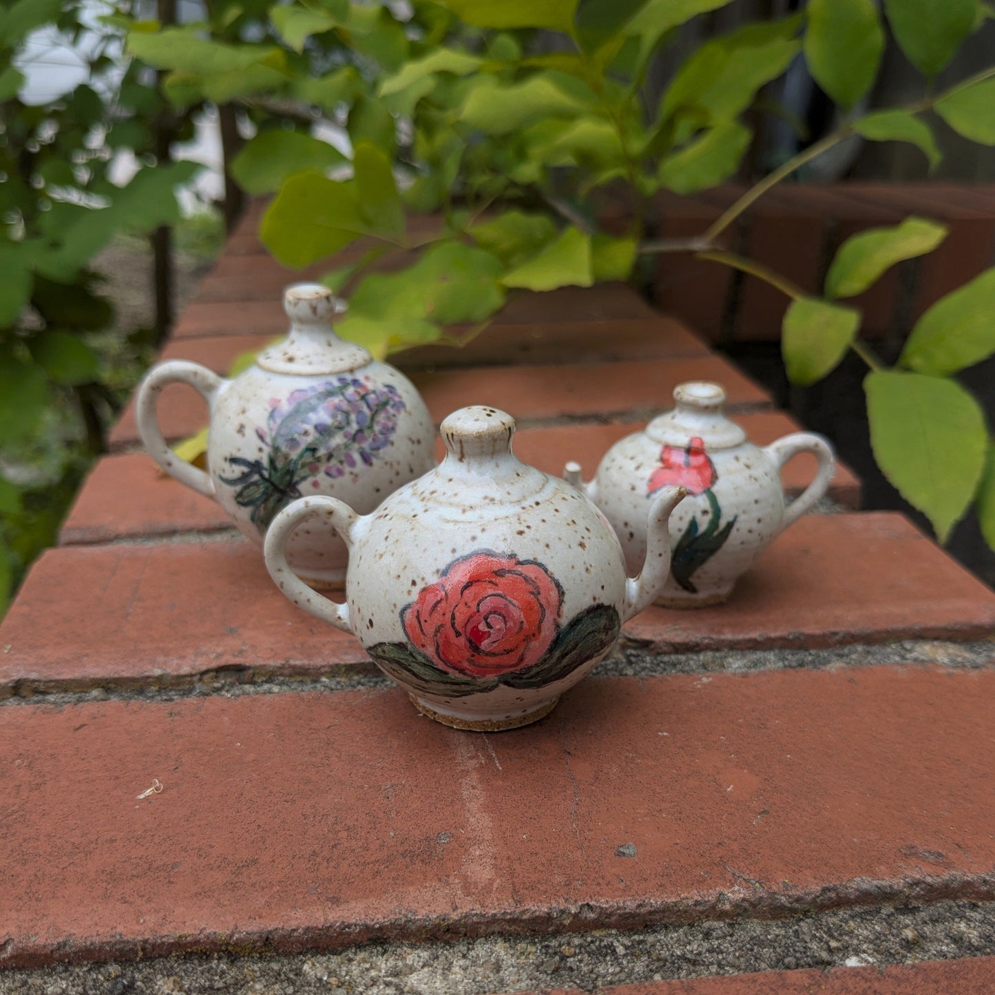 Three small ceramic teapots with floral designs on a brick surface.
