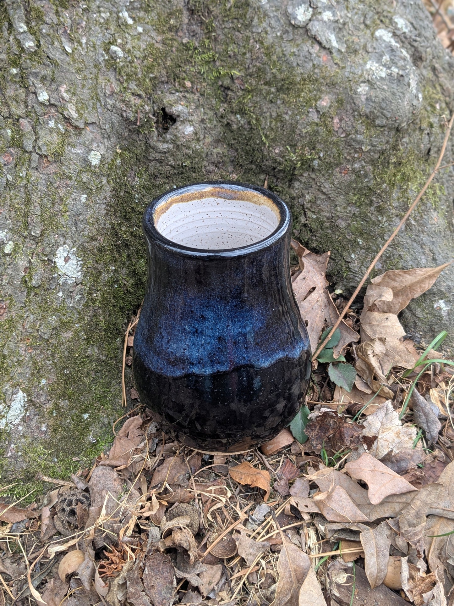 Ceramic vase with blue and black glaze on a natural background of leaves and tree bark.