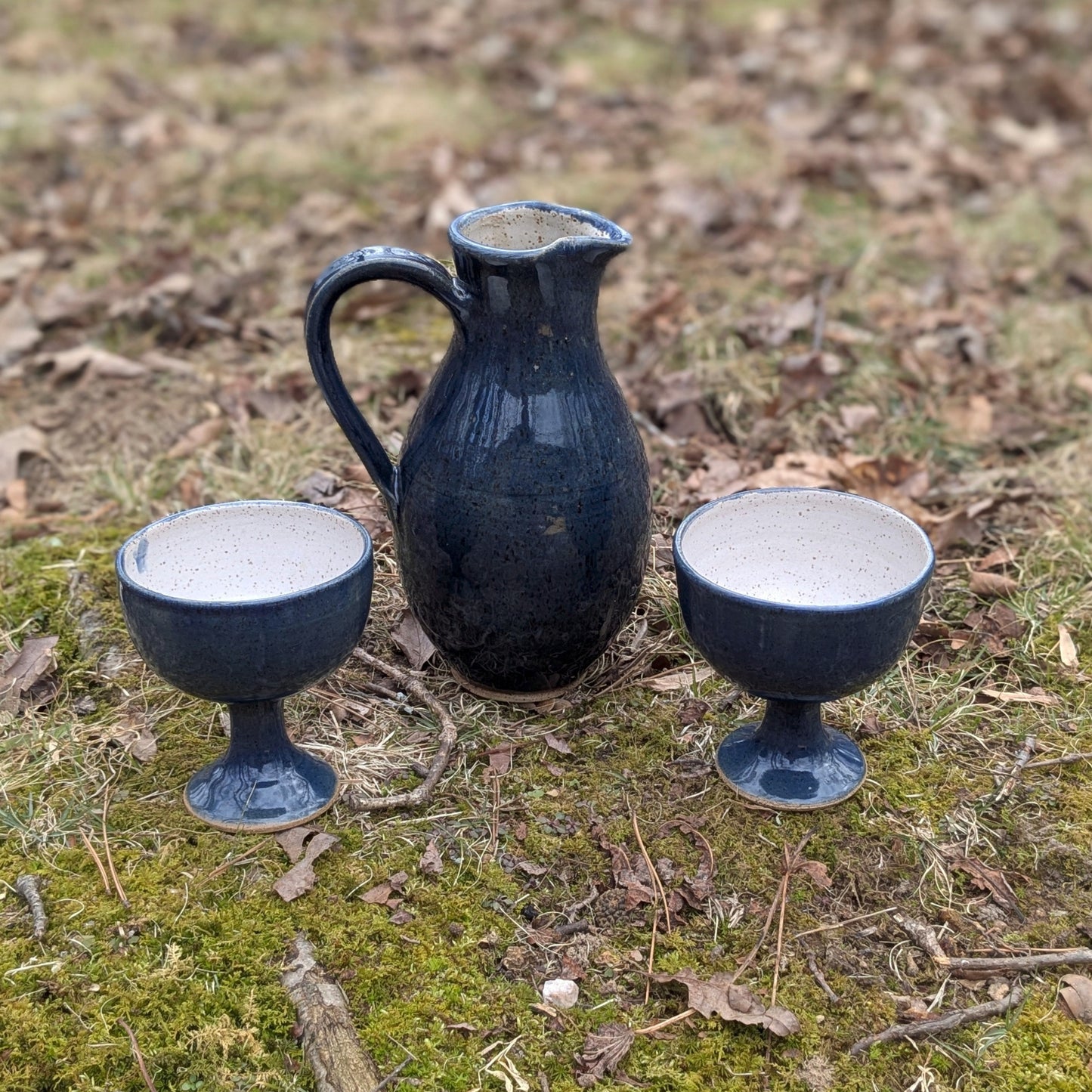 Two dark blue ceramic cups and a pitcher on a grassy ground with scattered leaves.