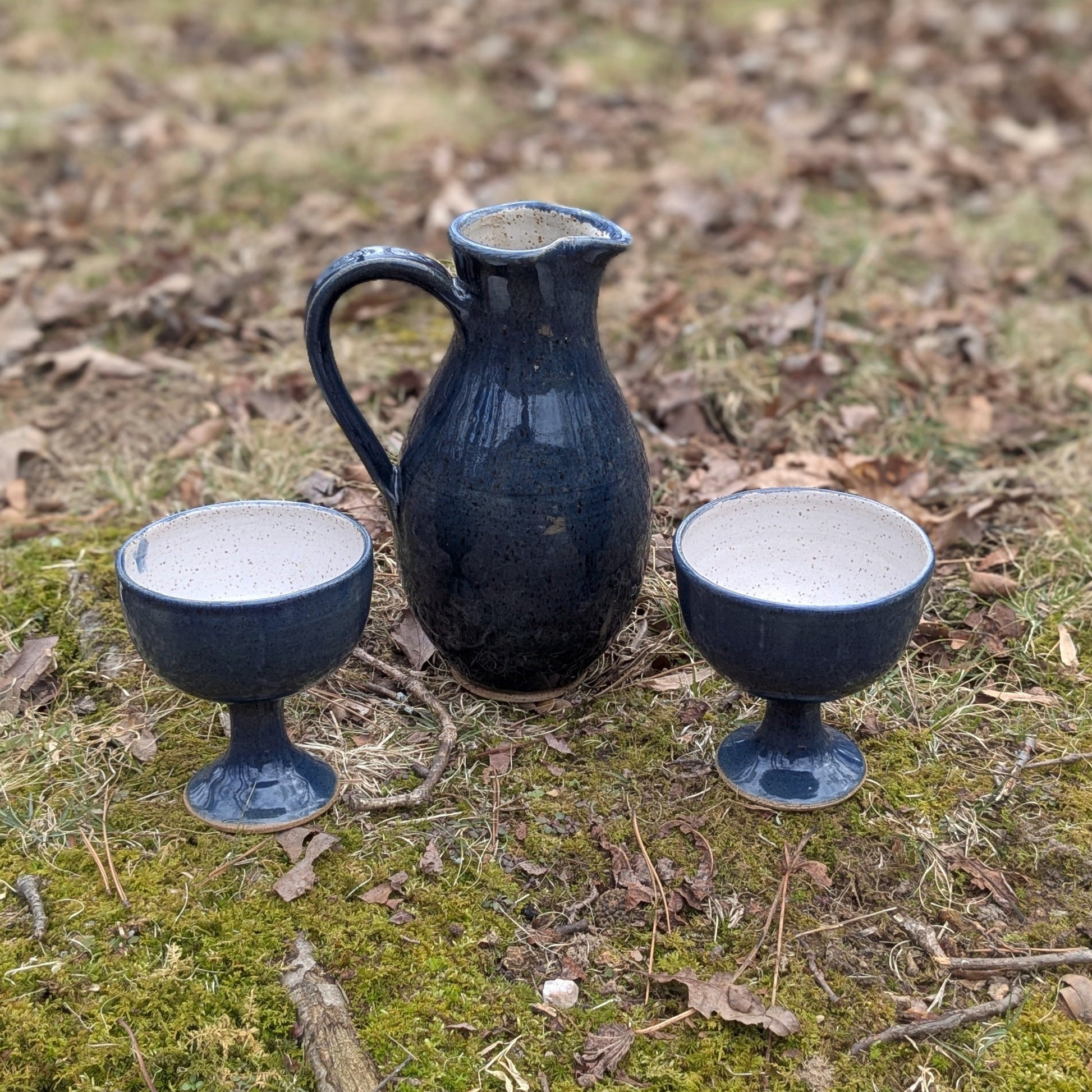 Two dark blue ceramic cups and a pitcher on a grassy ground with scattered leaves.